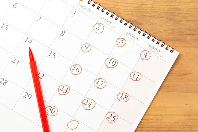 木製の机の上に置かれた卓上カレンダーと赤色のペンが写っています。カレンダーには複数の日付が赤丸で囲まれており、重要な予定や日程が示されているようです。