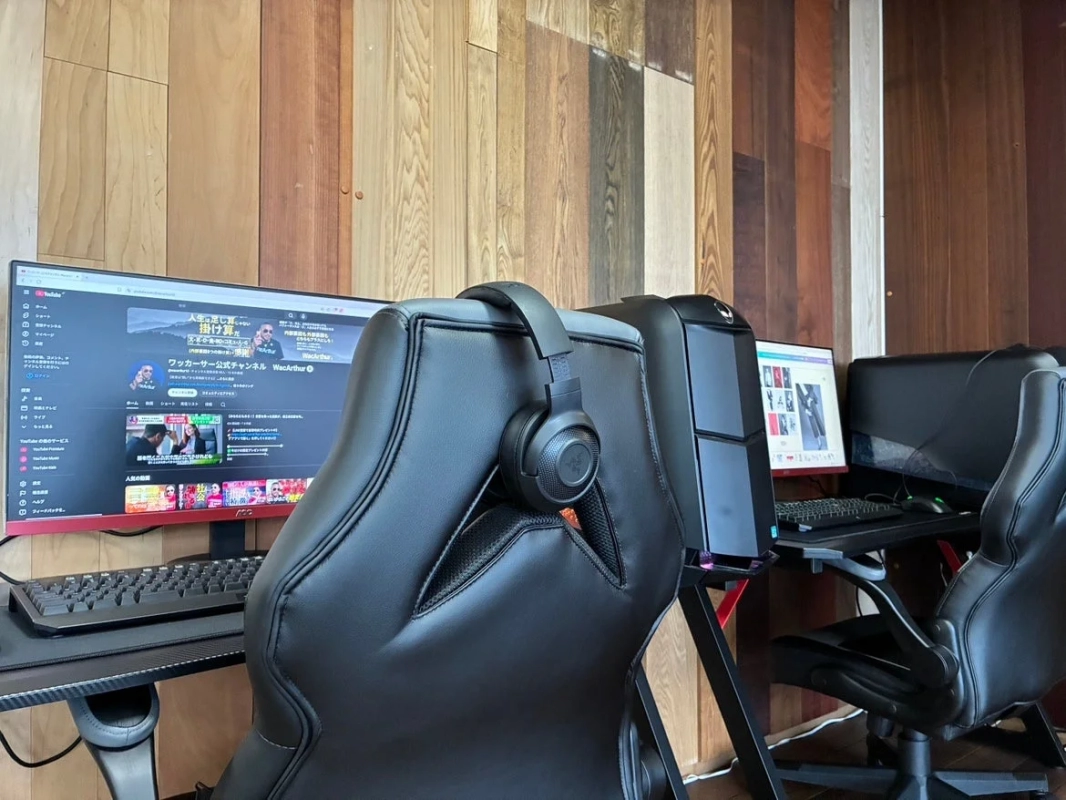 木目調の壁を背景に、ゲーミングPCと複数のモニター、黒いゲーミングチェアが設置された作業スペースの様子。左側のモニターにはYouTubeの画面が表示されており、高性能なヘッドホンがチェアに掛かっている。快適なPC環境がうかがえる。