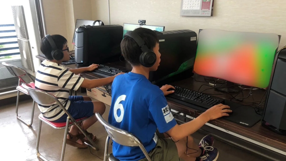 二人の少年がヘッドホンを装着し、各自のゲーミングPCに向かって真剣にゲームをプレイしている様子です。部屋には複数のPCが設置されています。