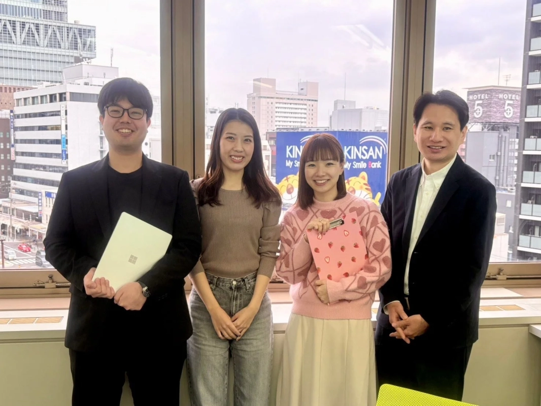 オフィスのような場所で、窓を背景に4人の男女が並んで笑顔で立っています。手前にはノートパソコンを持った男性もいます。