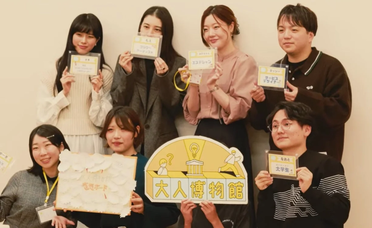 「大人博物館」と書かれたボードや、各自の氏名と職業が記されたカードを掲げる男女8人の集合写真
