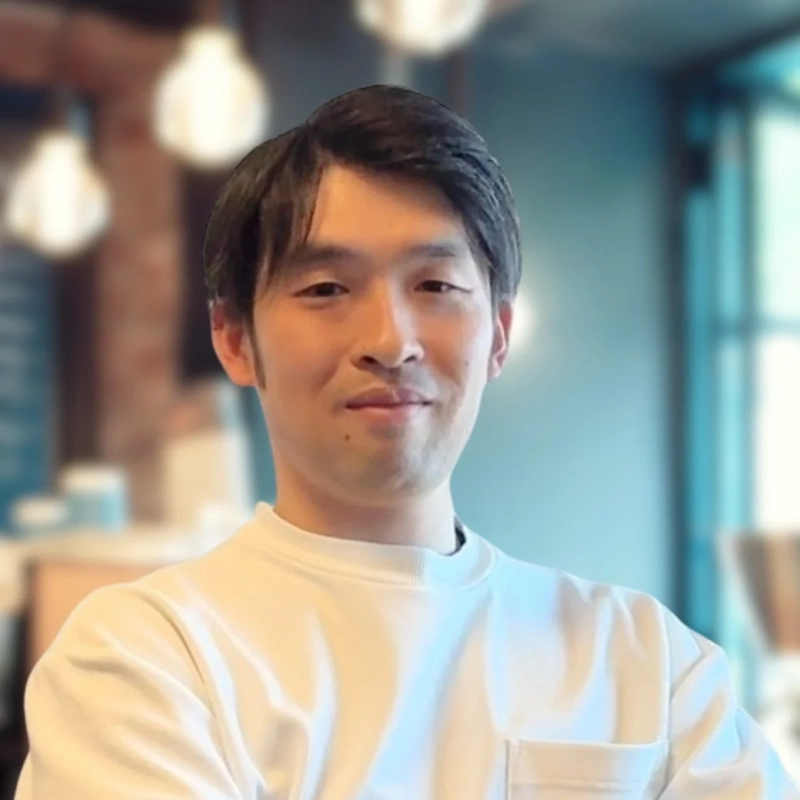 白いシャツを着たアジア人男性のポートレート。男性はわずかに微笑んでおり、背景はぼかされた屋内の空間で、いくつかの照明器具が見えます。