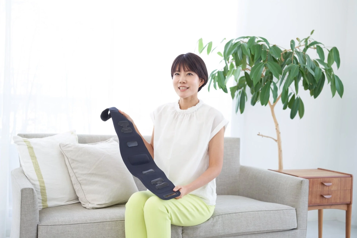 明るいリビングのソファに座った日本人女性が、健康や美容に使うと思われる黒いデバイスを持ち、笑顔を見せている