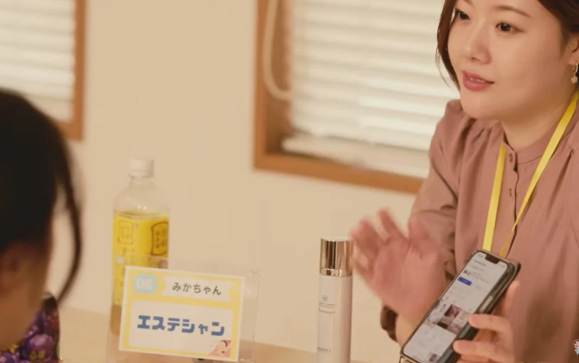 女性がスマートフォンを持ち、テーブルの上の名札（エステシャン、みかちゃん）や美容製品を前に、誰かに説明している様子