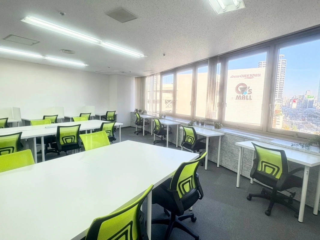 明るい日差しが差し込む窓際に、多くのデスクと鮮やかなライムグリーンの椅子が並べられた広々としたオフィススペースです。窓からは街の景色が見えます。