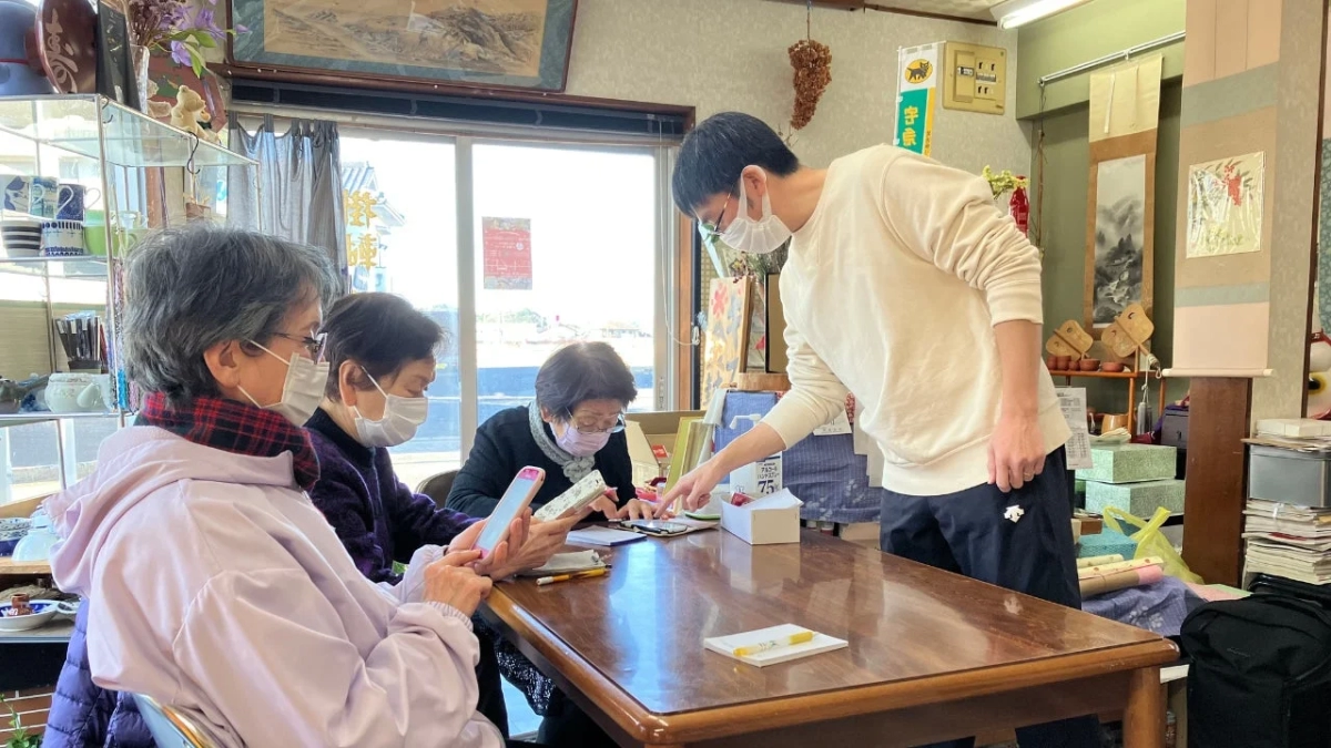 マスクを着用した若い男性が高齢の女性たちにスマートフォンの使い方を教えている様子。伝統的な雰囲気の店内で、デジタル機器の学習会が行われている。
