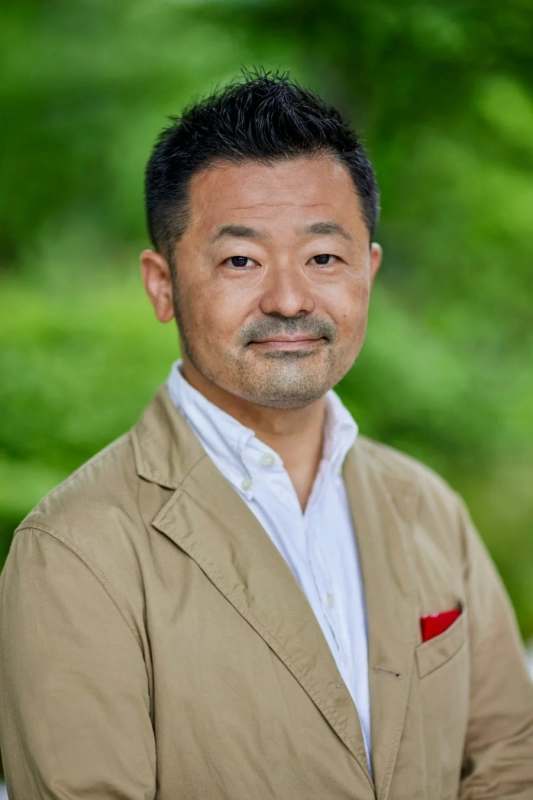 ベージュのジャケットを着用したアジア人男性が、緑のぼかし背景の前でカメラに視線を向けているポートレート写真です。