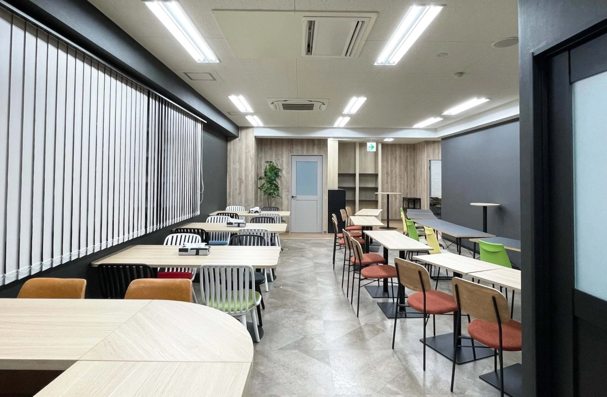 明るい照明とモダンな内装が特徴のオフィス共有スペース。木目調のテーブルとカラフルな椅子が多数配置されており、休憩や作業に適した開放的な空間です。